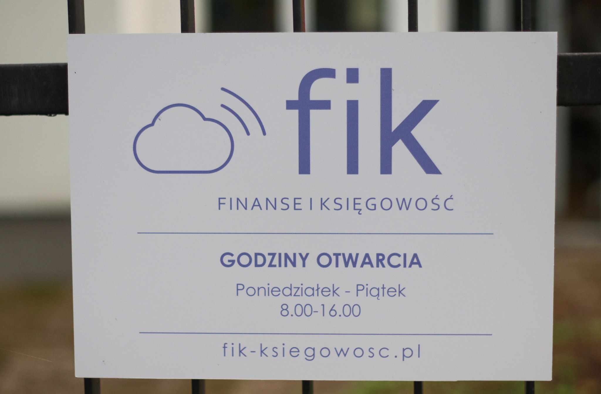 Tablica informacyjna firmy Fik Finanse i Księgowość w Gdyni, z logo chmury, godzinami otwarcia i adresem strony internetowej, umieszczona na tle metalowego ogrodzenia.