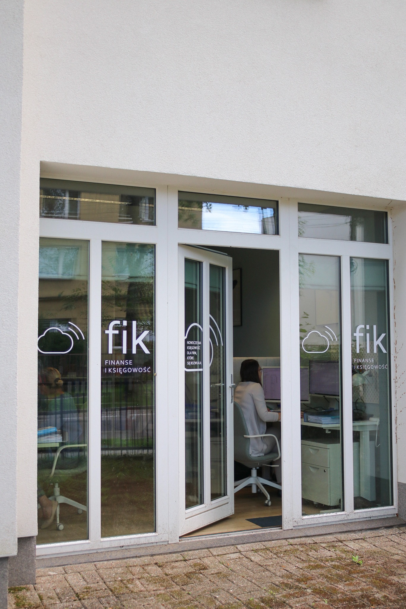 Wejście do biura 'fik Finanse i Księgowość' z otwartymi drzwiami, przez które widać pracującą kobietę przy biurku z komputerami. Na szybach logo firmy.