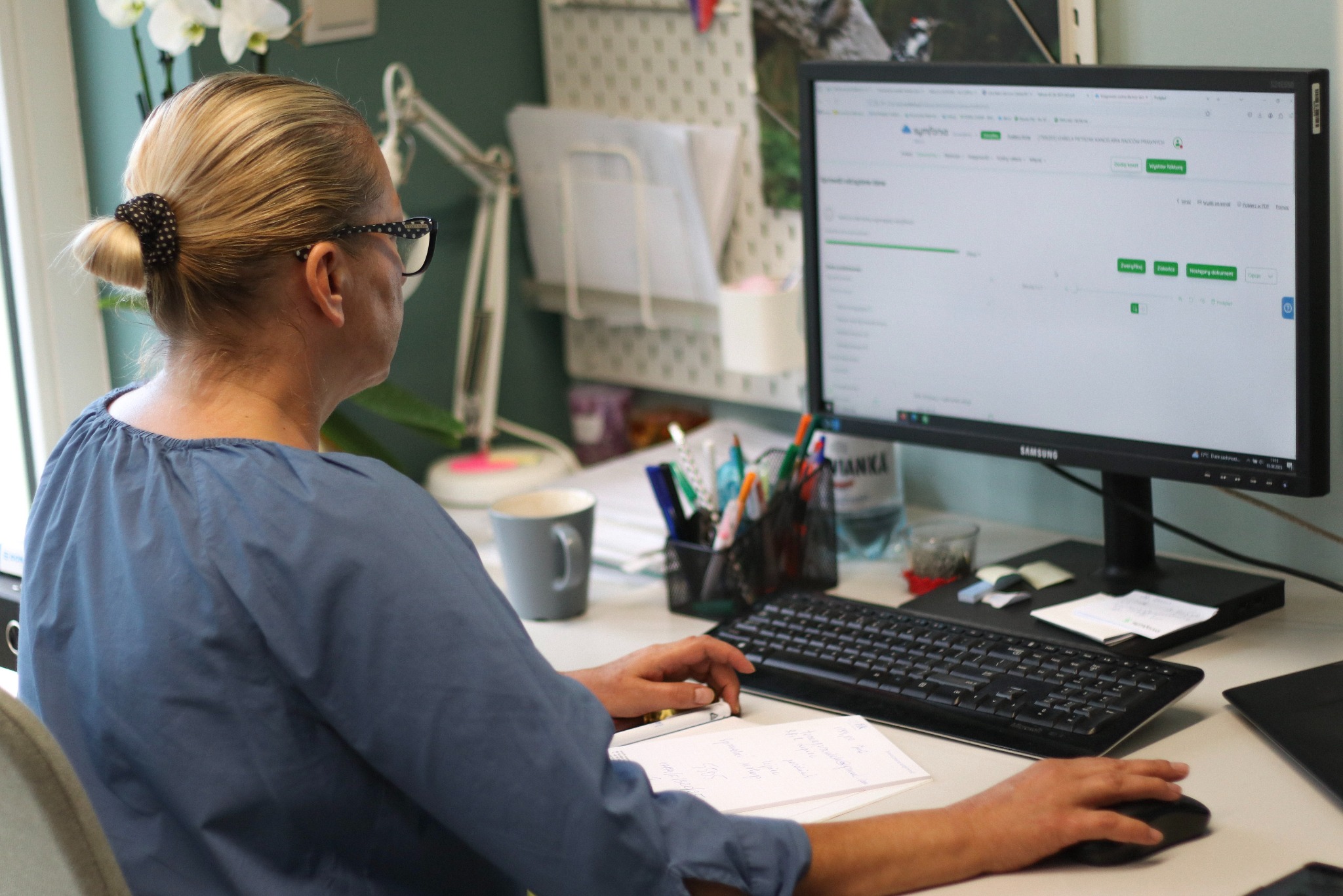 Kobieta w okularach pracuje przy biurku z komputerem, notatkami i przyborami. Widok zza pleców, monitor wyświetla program księgowy. Jasne biuro, kwiaty w tle.