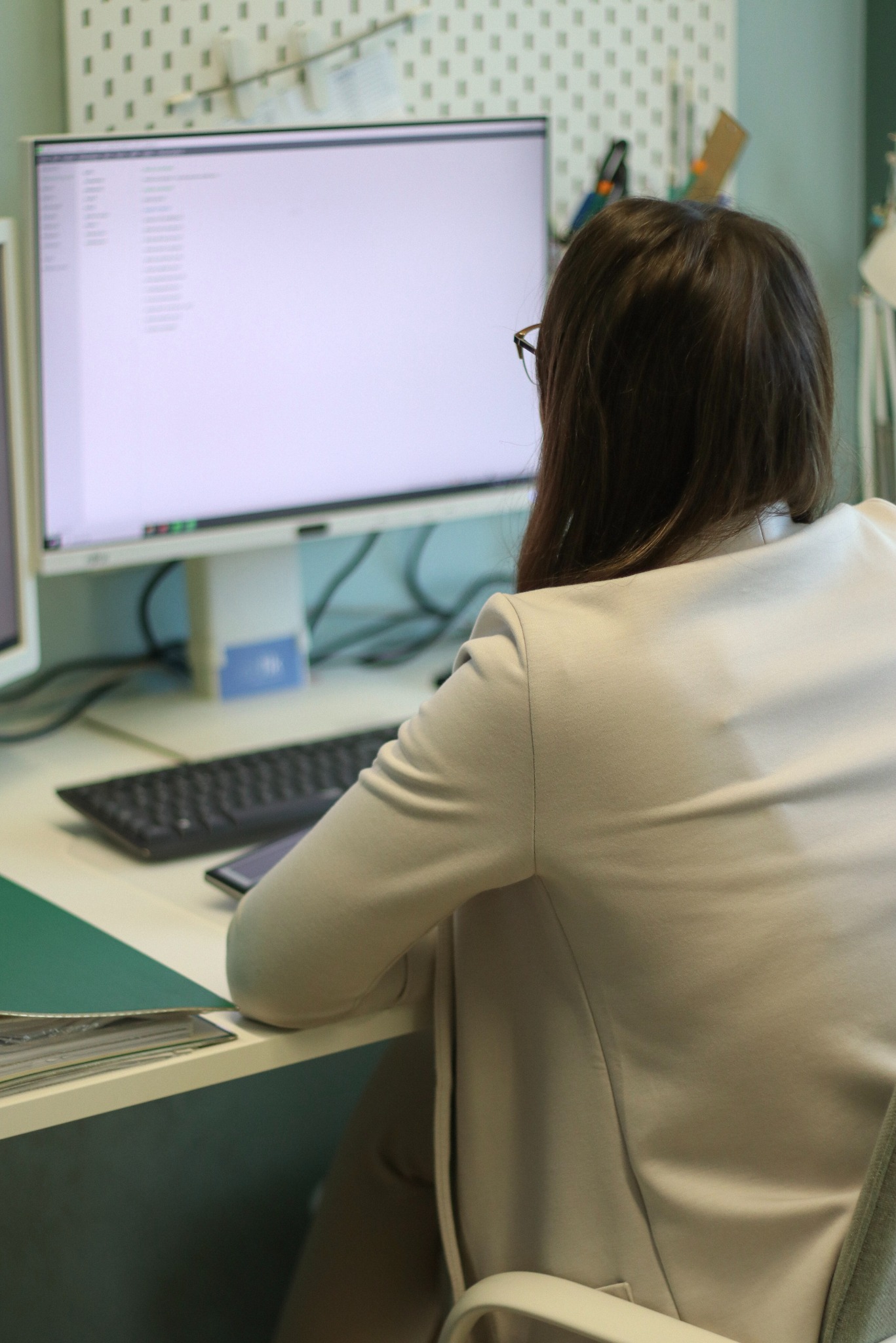 Kobieta w jasnym żakiecie siedzi przy biurku przed monitorem z listą. Widok z tyłu, skupienie na monitorze i dokumentach na biurku.