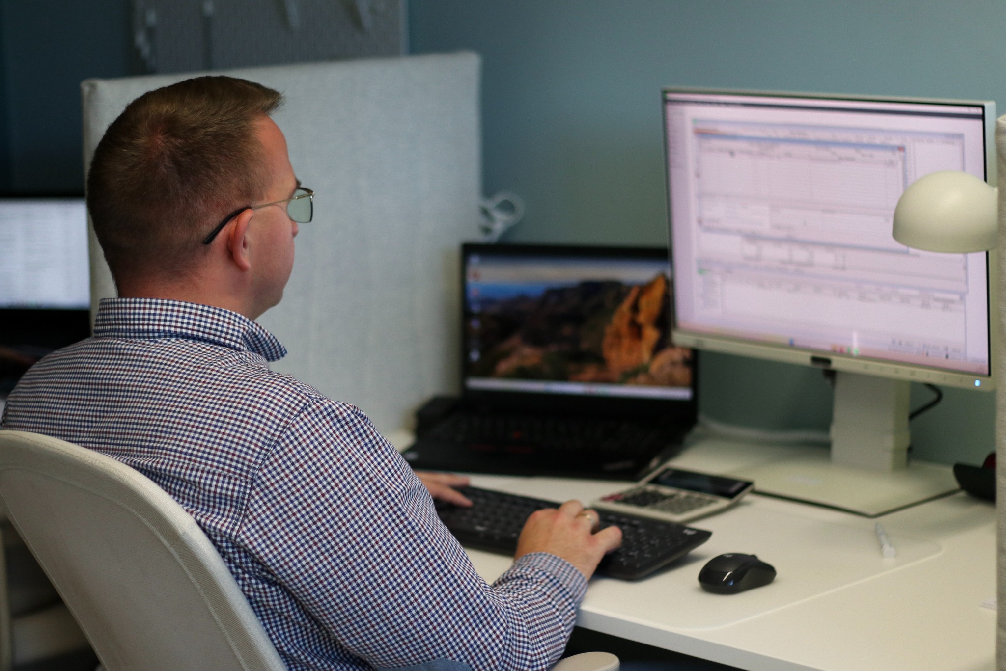 Księgowy w okularach, tyłem, pracuje przy biurku z komputerem i kalkulatorem. Widoczny arkusz kalkulacyjny na monitorze.