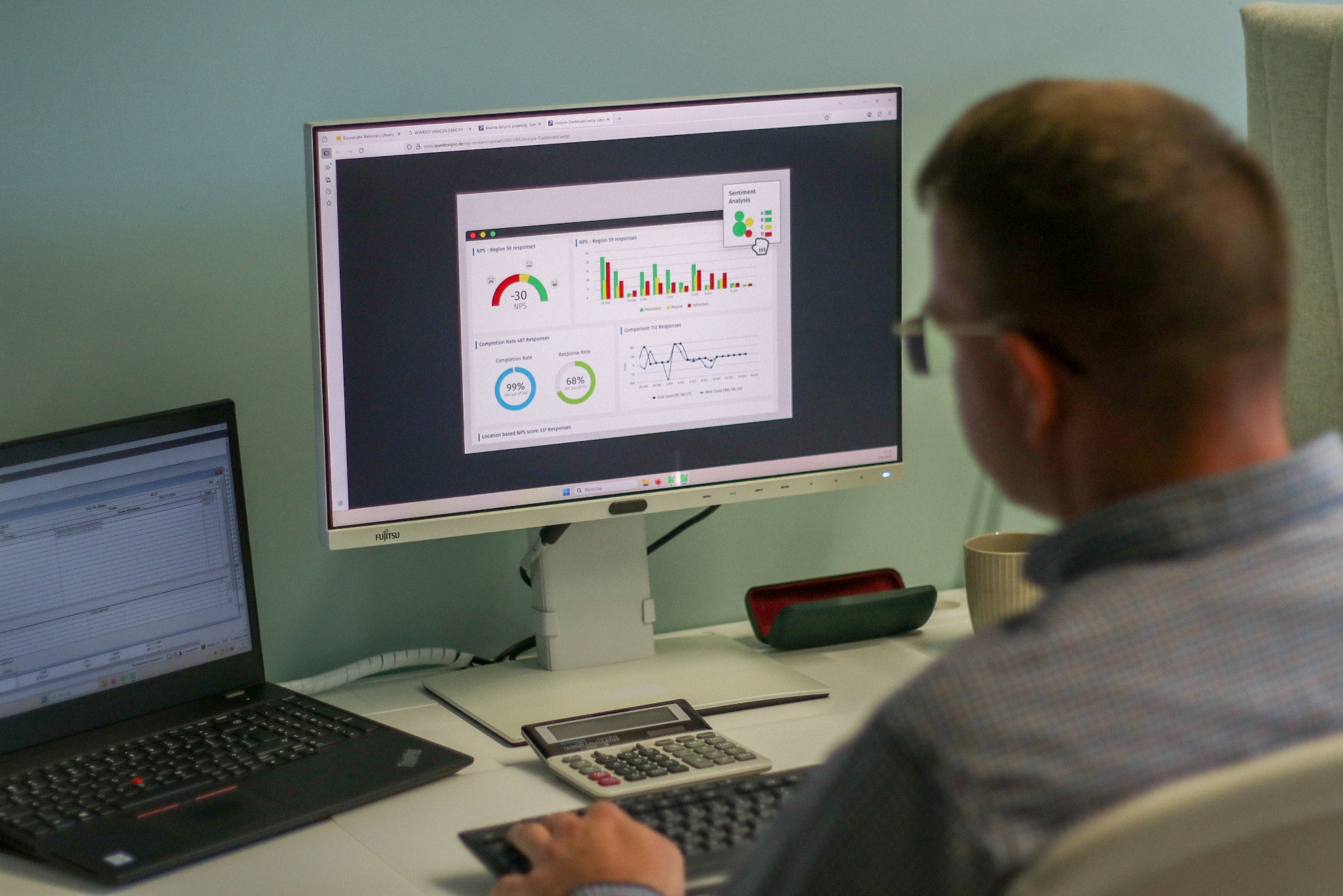 Mężczyzna analizuje dane na monitorze Fujitsu, obok laptop Lenovo, kalkulator i kubek. Widoczne wykresy NPS i analizy sentymentu. Praca biurowa.