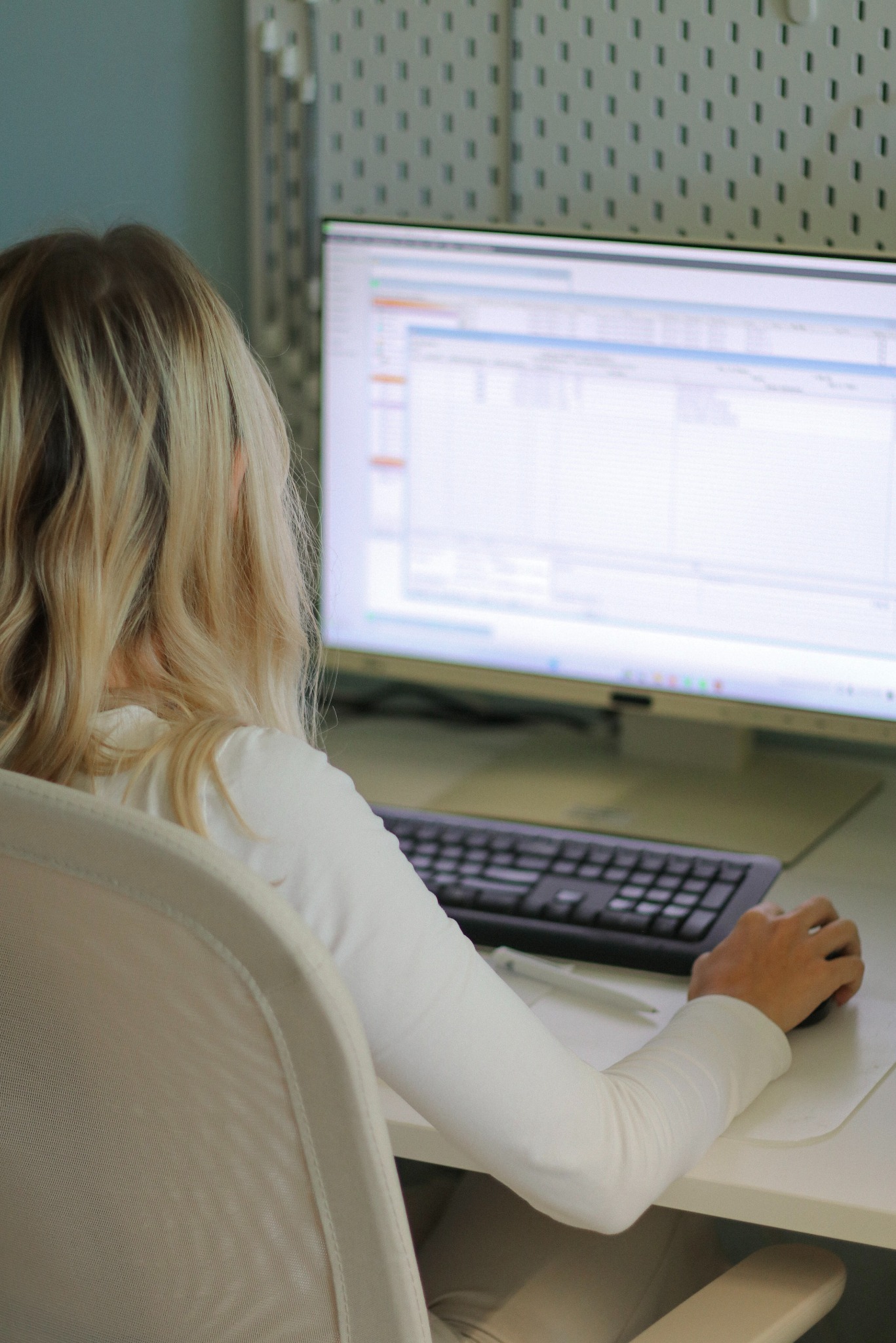 Kobieta z blond włosami siedzi przy biurku, używa myszki i patrzy na monitor z arkuszem kalkulacyjnym. Na biurku leży długopis.