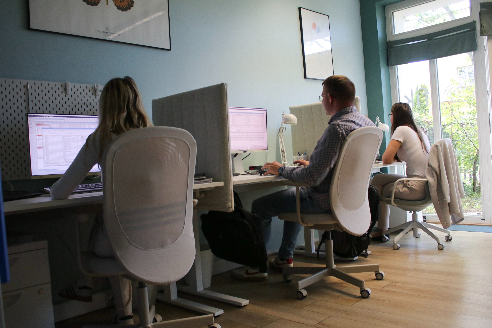 Jasne biuro z trzema osobami pracującymi przy biurkach. Monitory z danymi, nowoczesne krzesła i naturalne światło z okna tworzą profesjonalne środowisko pracy.