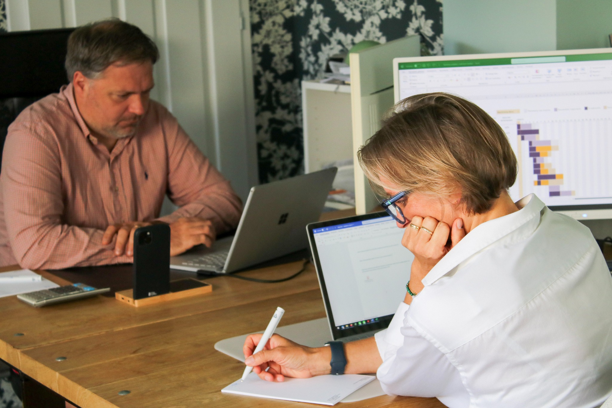 Dwie osoby pracują przy biurku: kobieta pisze, opierając głowę na ręce, a mężczyzna korzysta z laptopa. W tle widoczny monitor z wykresem Gantta.