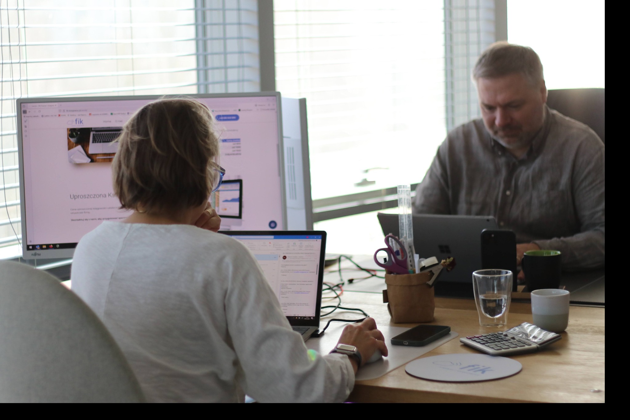 Dwie osoby pracują w biurze przy komputerach. Na biurku kalkulator, szklanka wody i przybory biurowe. W tle okno z żaluzjami i logo firmy na monitorze.