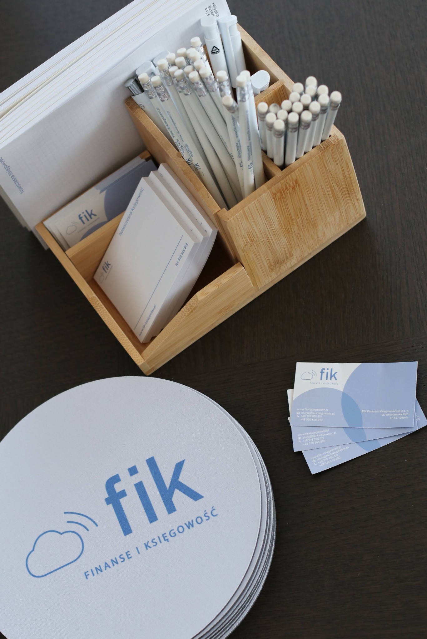 Biurko z drewnianym organizerem wypełnionym notatnikami, ołówkami i wizytówkami firmy Fik Finanse i Księgowość. Na blacie leżą podkładki z logo firmy.