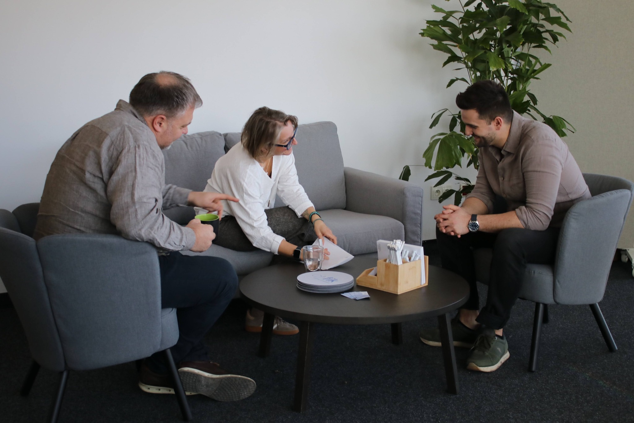 Trzy osoby w swobodnej atmosferze biura omawiają dokumenty przy okrągłym stoliku. W tle szara sofa, krzesła i roślina doniczkowa. Jasne wnętrze.