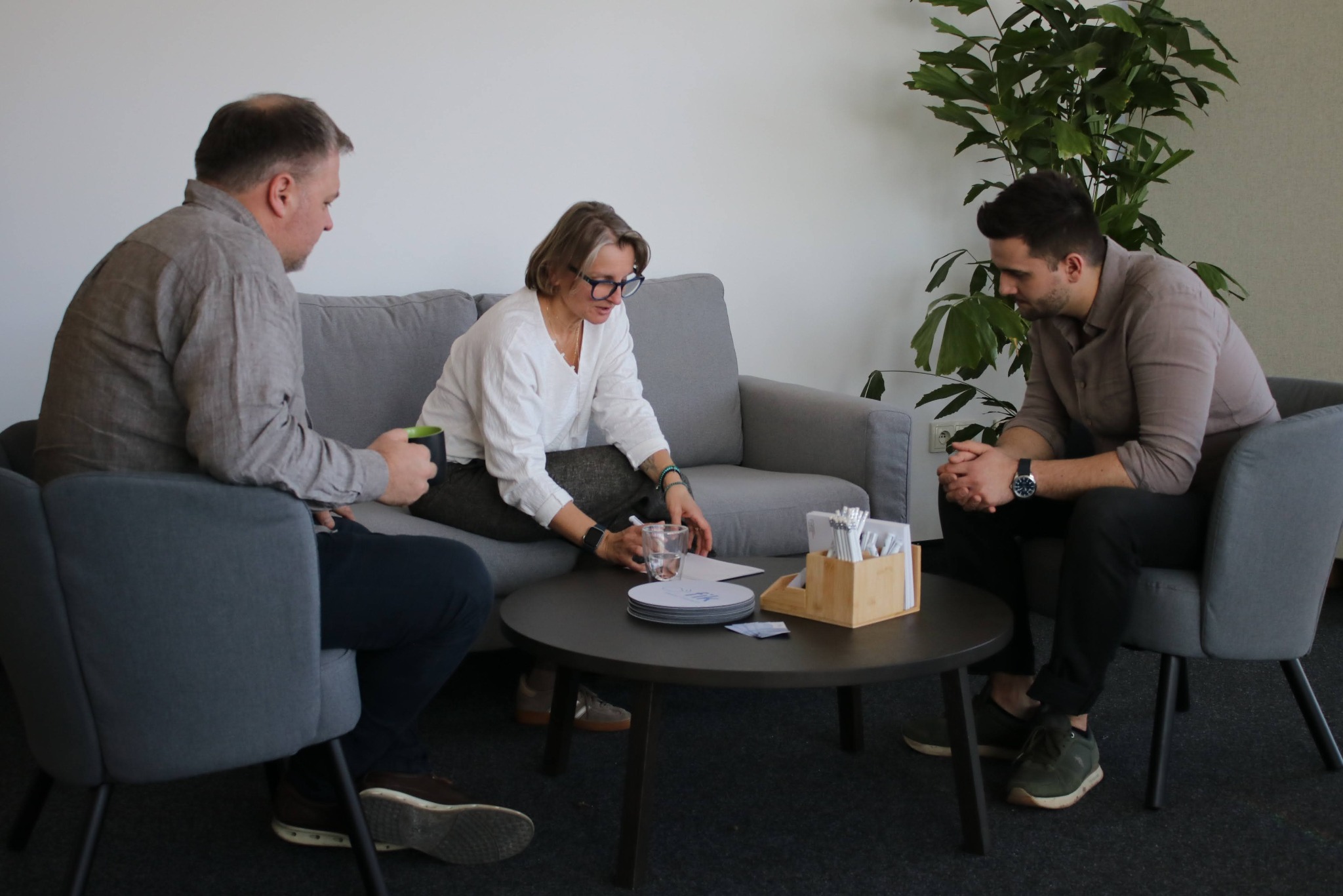 Spotkanie biznesowe w jasnym wnętrzu: trzech konsultantów omawia dokumenty przy okrągłym stole. Minimalistyczny wystrój, roślina doniczkowa w tle.