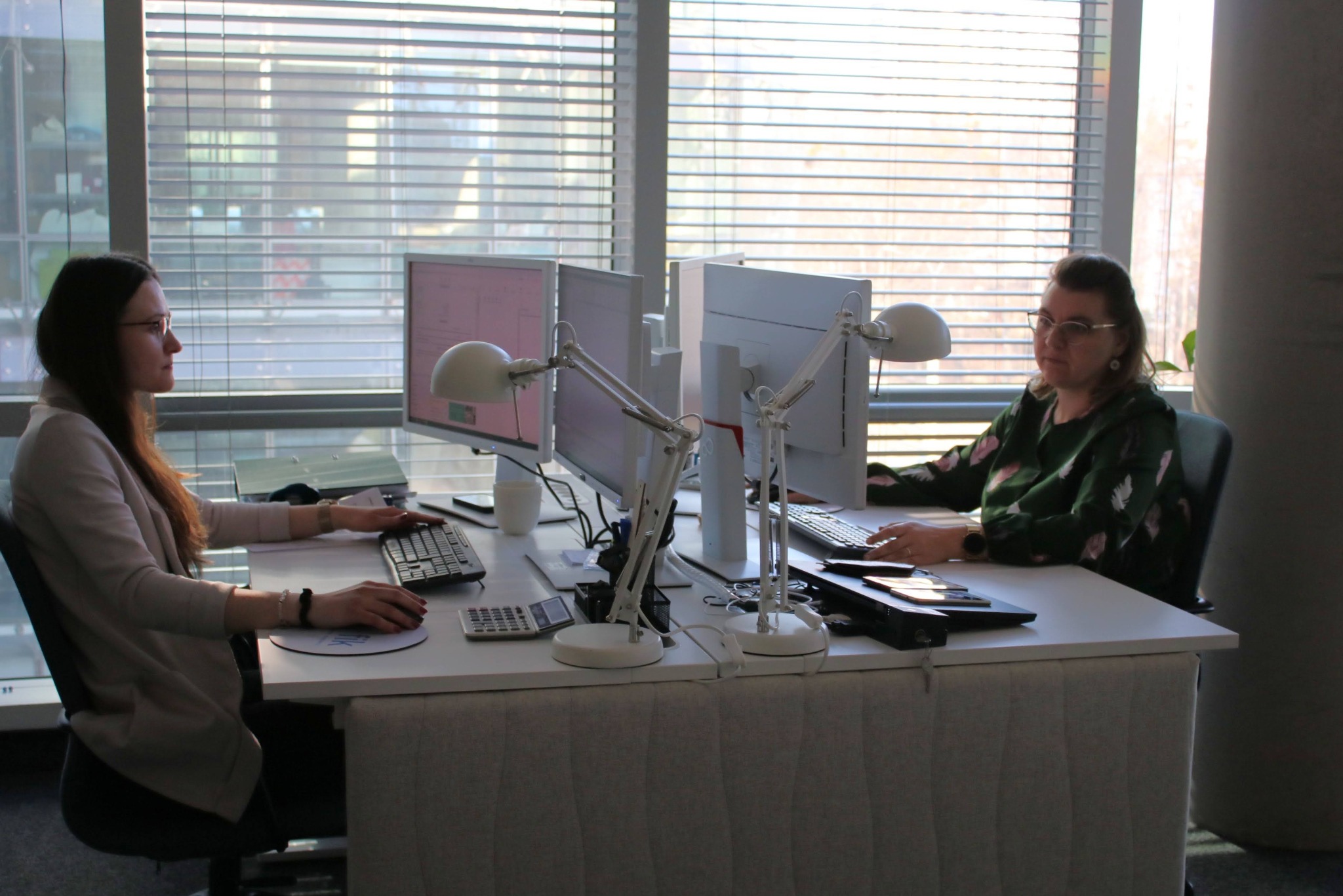 Dwie kobiety pracują przy biurkach z monitorami i lampkami w jasnym biurze z żaluzjami. Widoczne kalkulatory i akcesoria biurowe. Ujęcie z perspektywy.