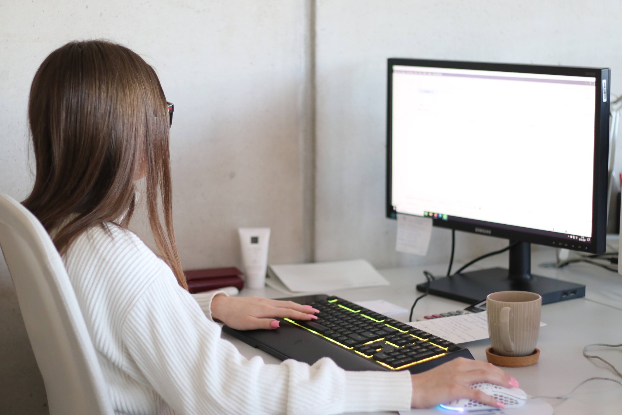 Kobieta w swetrze przy biurku z podświetlaną klawiaturą i monitorem. Widok zza pleców, skupienie na dłoniach na klawiaturze i myszce. Kubek z napojem na biurku.