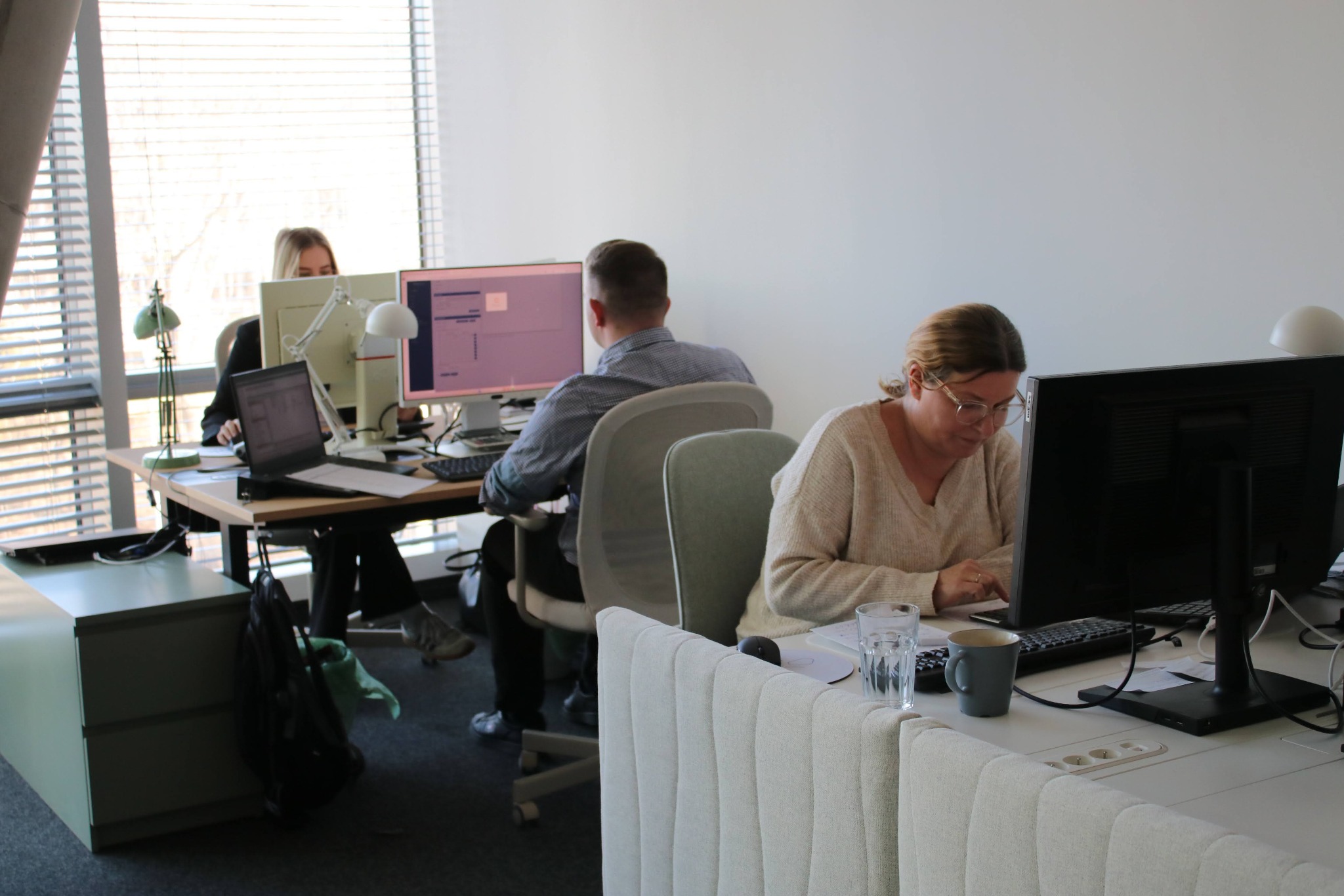 Jasne biuro: trzy osoby pracują przy biurkach z komputerami. Widoczne monitory, laptopy, lampki i akcesoria biurowe. Duże okno z żaluzjami w tle.