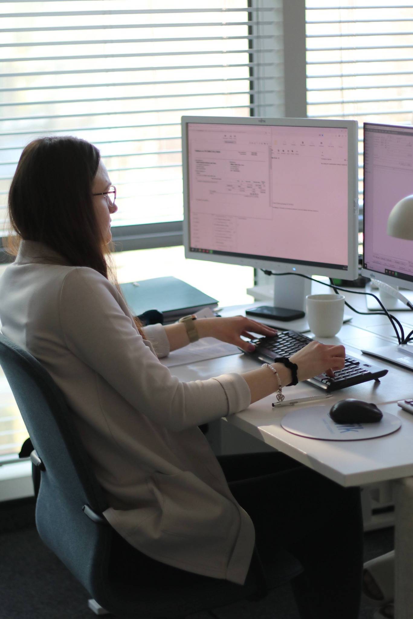 Kobieta w okularach pisze na klawiaturze przed monitorem w biurze. Na biurku myszka, podkładka, długopis i kubek. W tle żaluzje okienne.