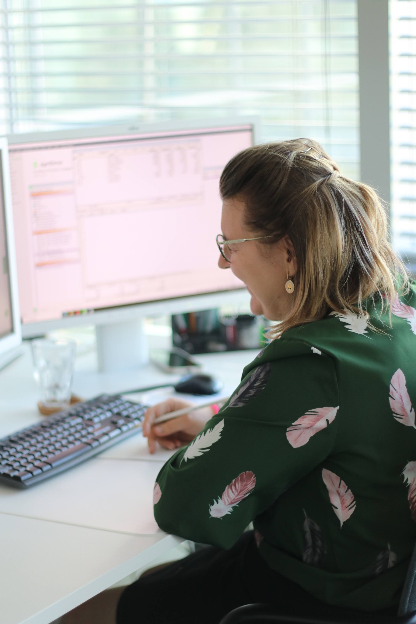 Kobieta w zielonej bluzce z piórami pracuje przy biurku z dwoma monitorami. Widoczna klawiatura, szklanka i długopis. Jasne biuro z żaluzjami w tle.