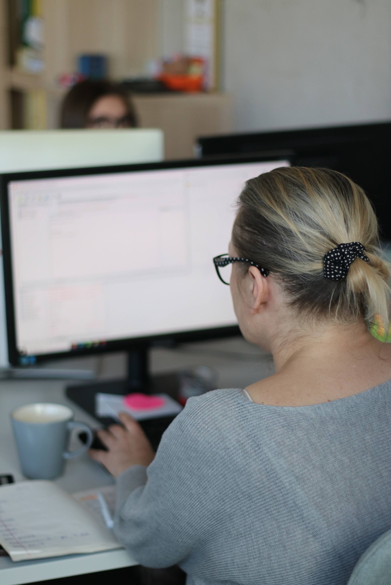 Pracownica biura w szarym swetrze, z włosami spiętymi w kucyk, pracuje przy komputerze. Na biurku notatnik, kubek i myszka. W tle inna osoba przy stanowisku pracy.