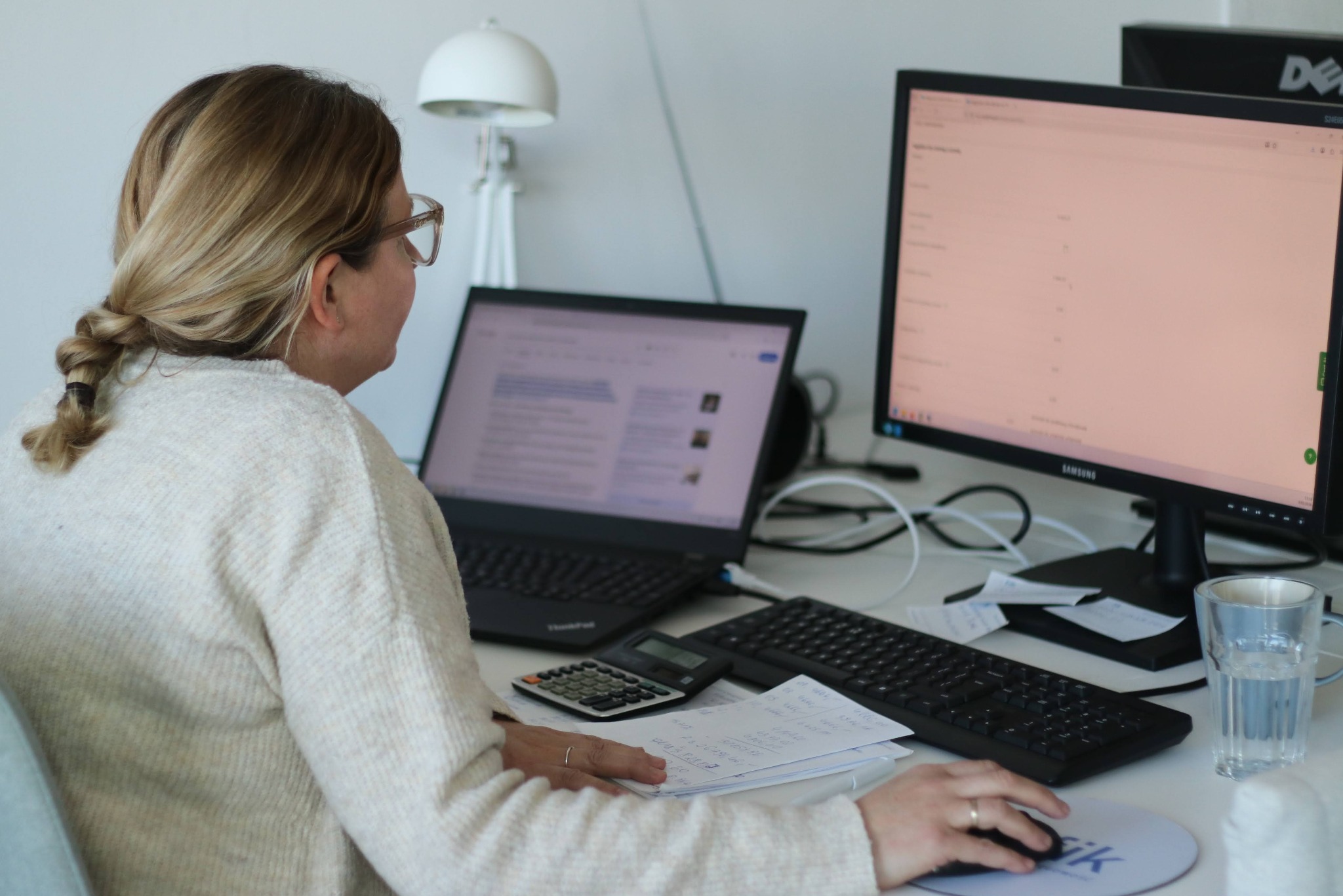 Kobieta w biurze, z laptopem, monitorem, kalkulatorem i dokumentami na biurku. Praca biurowa, analiza danych, obliczenia. Jasne, naturalne oświetlenie.