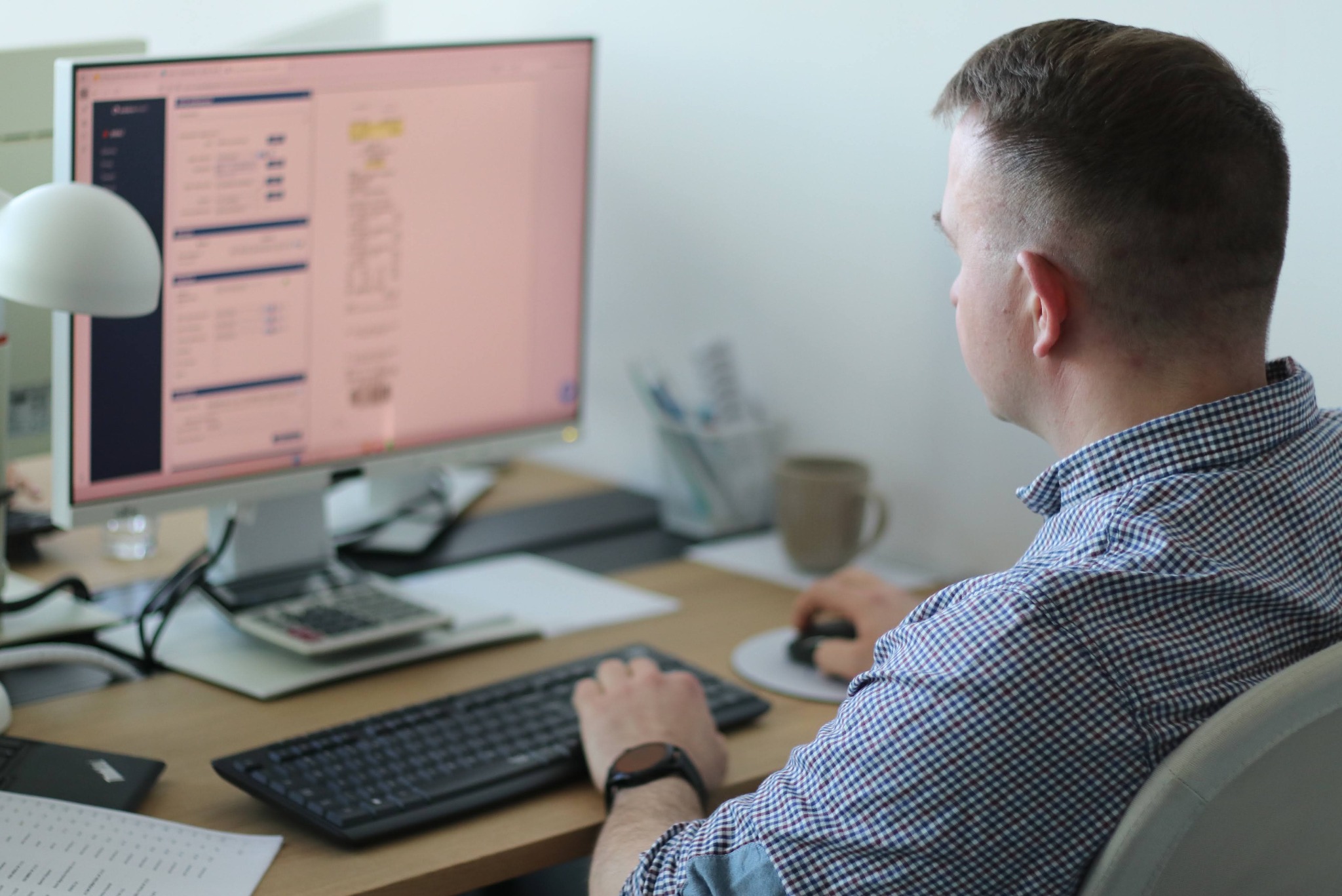 Mężczyzna w koszuli w kratę pracuje przy biurku z komputerem, klawiaturą i kalkulatorem. Na ekranie widoczne dane tabelaryczne. Kubek i lampka na biurku.