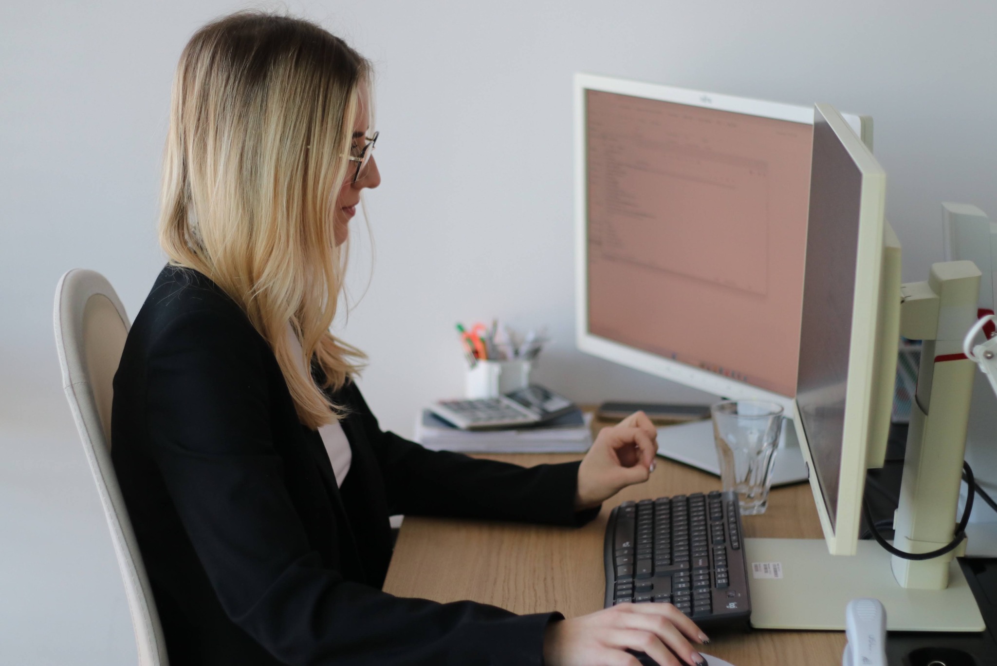 Kobieta w okularach i marynarce pracuje przy biurku z dwoma monitorami. Na biurku kalkulator, szklanka i przybory biurowe. Jasne tło, profil.