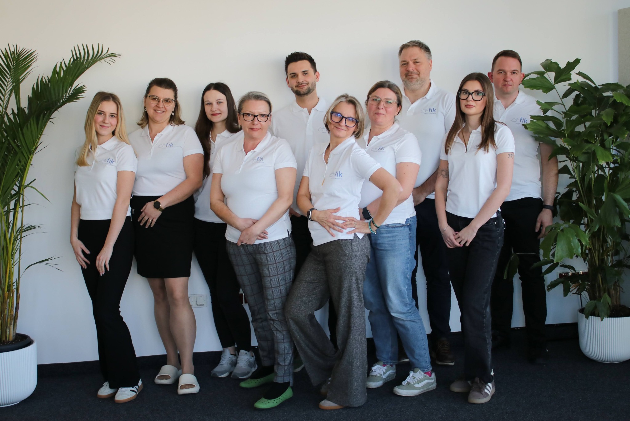 Zespół w białych koszulkach polo z logo firmy, stojący na tle białej ściany z roślinami doniczkowymi po bokach. Uśmiechnięte twarze, różnorodne stylizacje spodni.