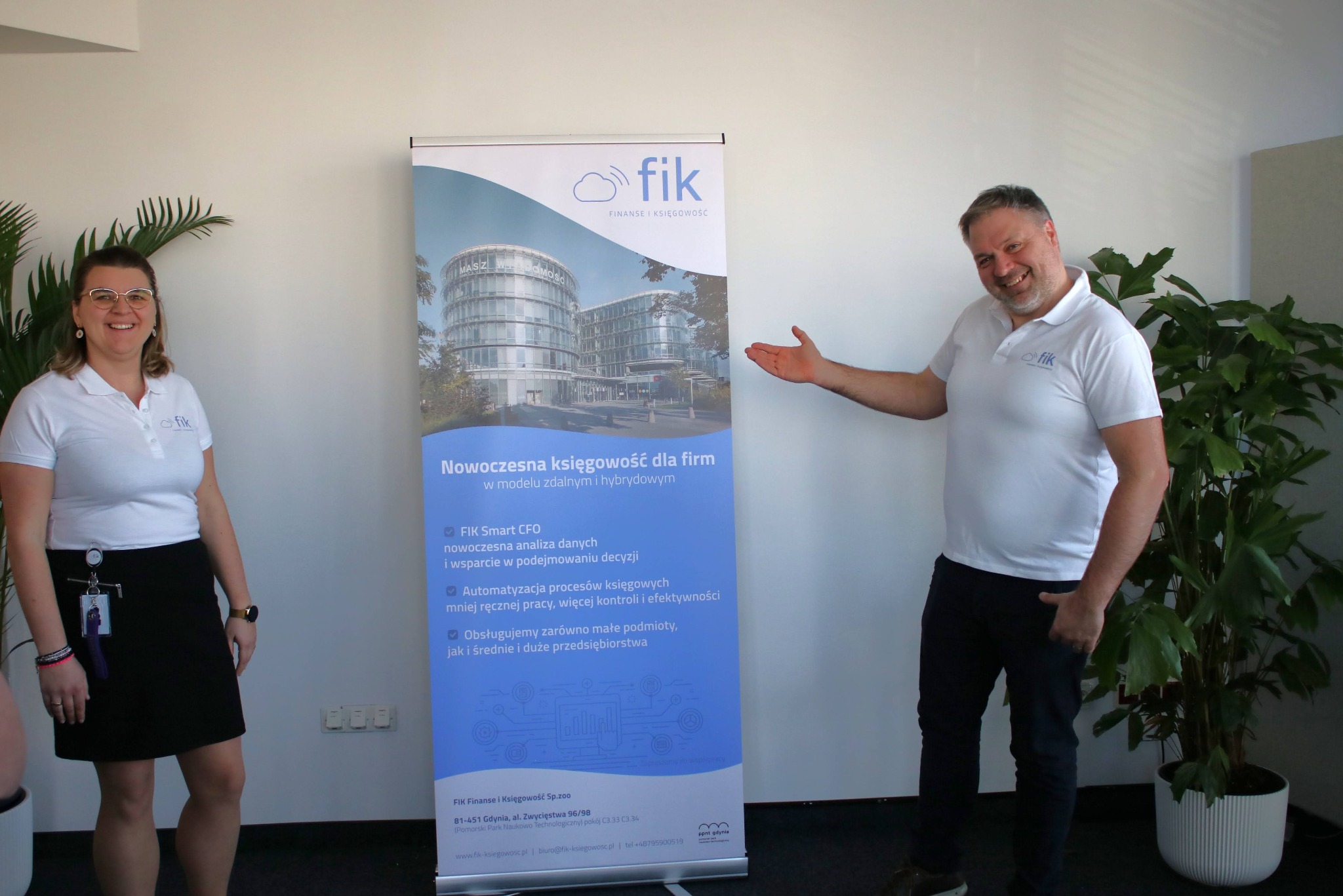 Dwie osoby prezentują roll-up z ofertą nowoczesnej księgowości firmy FIK. Na roll-upie budynek biurowy. Pracownicy w koszulkach polo z logo firmy. W tle biuro i roślina doniczkowa.