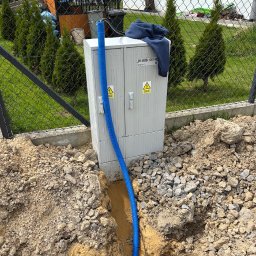 Adrian Siekierski - Szafka elektryczna ZK-BBB 120746 z niebieskim przewodem w wykopie, na tle zielonego trawnika i ogrodzenia z siatki, częściowo zasłonięta bluzą.