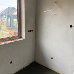 Siekierski Smart Elektryk - Surowy stan wnętrza po wylewce, z widocznymi puszkami elektrycznymi w ścianie i wystającymi przewodami. W tle okno z widokiem na dom i maszynę budowlaną.