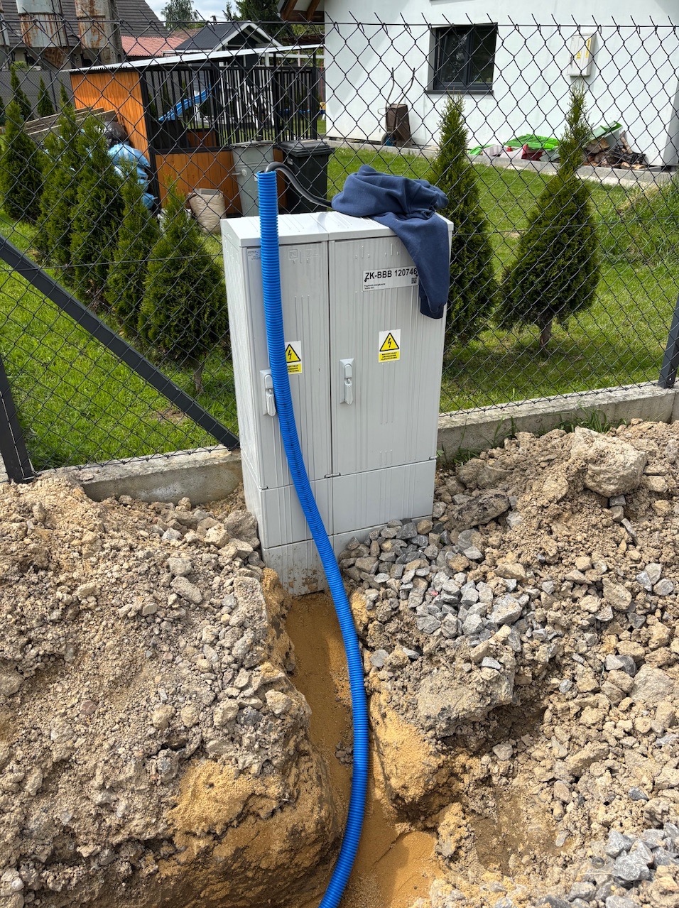 Szafka elektryczna ZK-BBB 120746 z niebieskim przewodem w wykopie, na tle zielonego trawnika i ogrodzenia z siatki, częściowo zasłonięta bluzą.