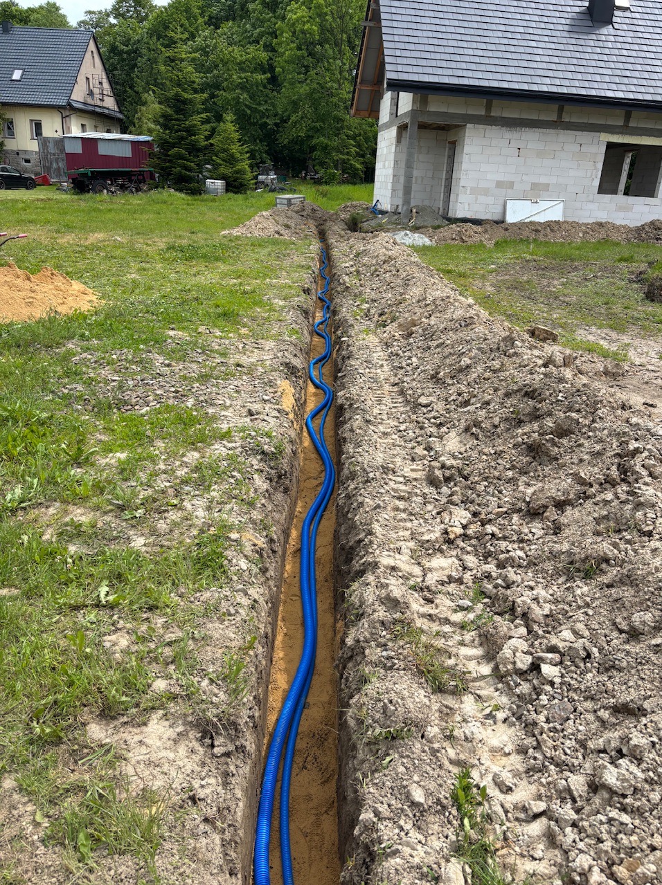 Dwa niebieskie kable w wykopanym rowie ziemnym, prawdopodobnie przygotowane do instalacji elektrycznej, z domem w tle w trakcie budowy.