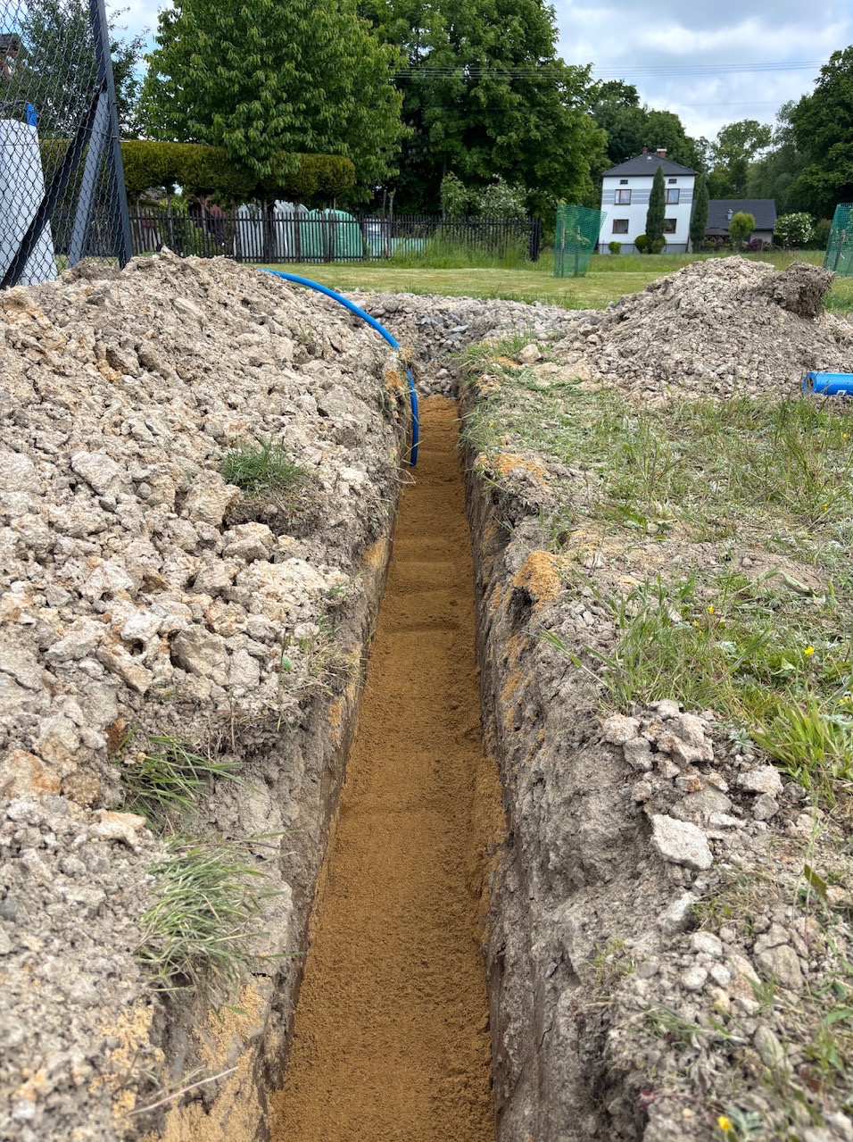 Wykop pod niebieski kabel w ziemi, Bielsko-Biała. Wykopana bruzda w trawie, przygotowanie do ułożenia instalacji. Widok na dom w tle, dzień.