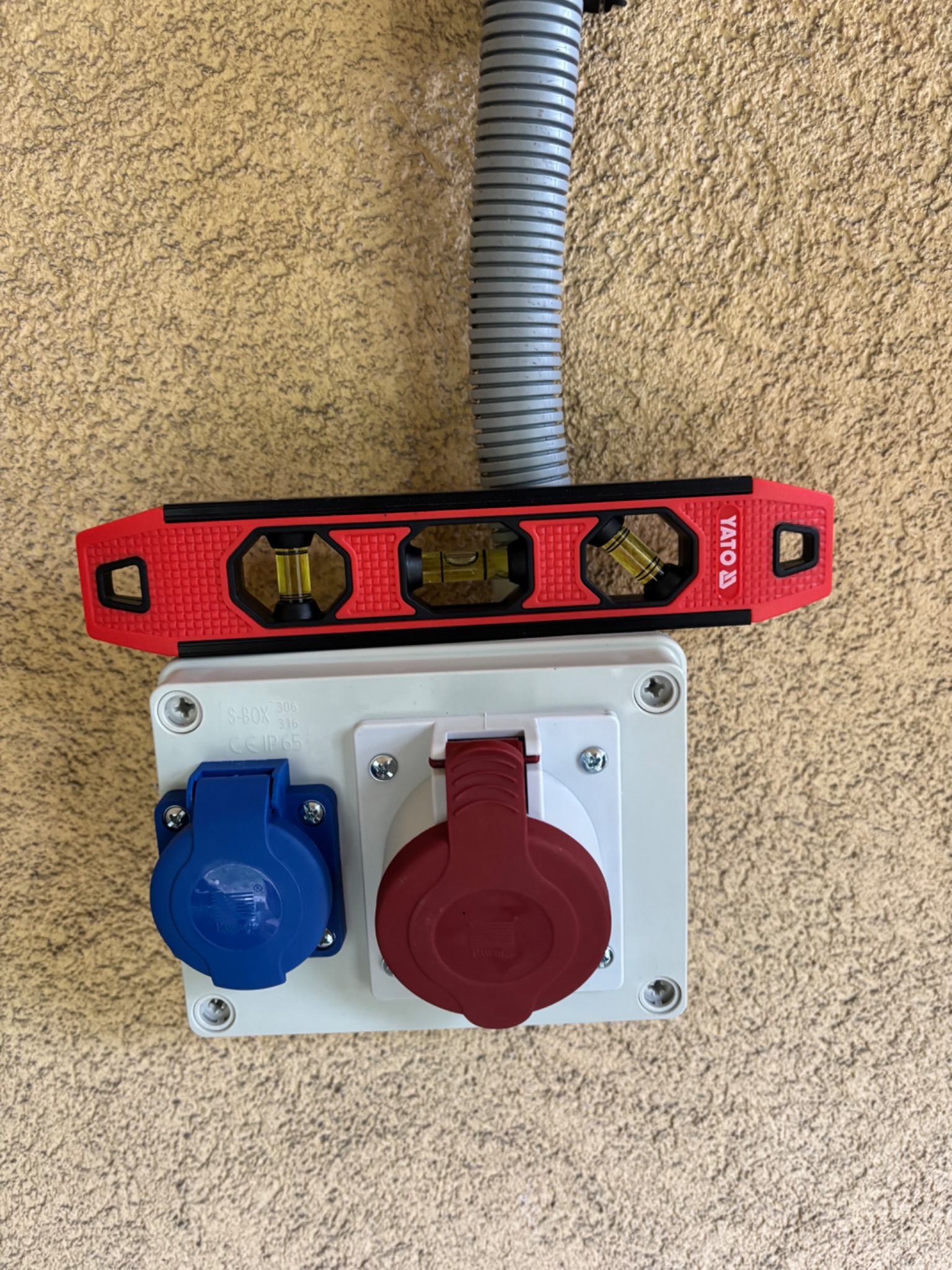 Gniazdo siłowe w białej obudowie z niebieskim i czerwonym gniazdem, poziomica Yato na górze, szara rura karbowana, beżowa ściana o nierównej fakturze.