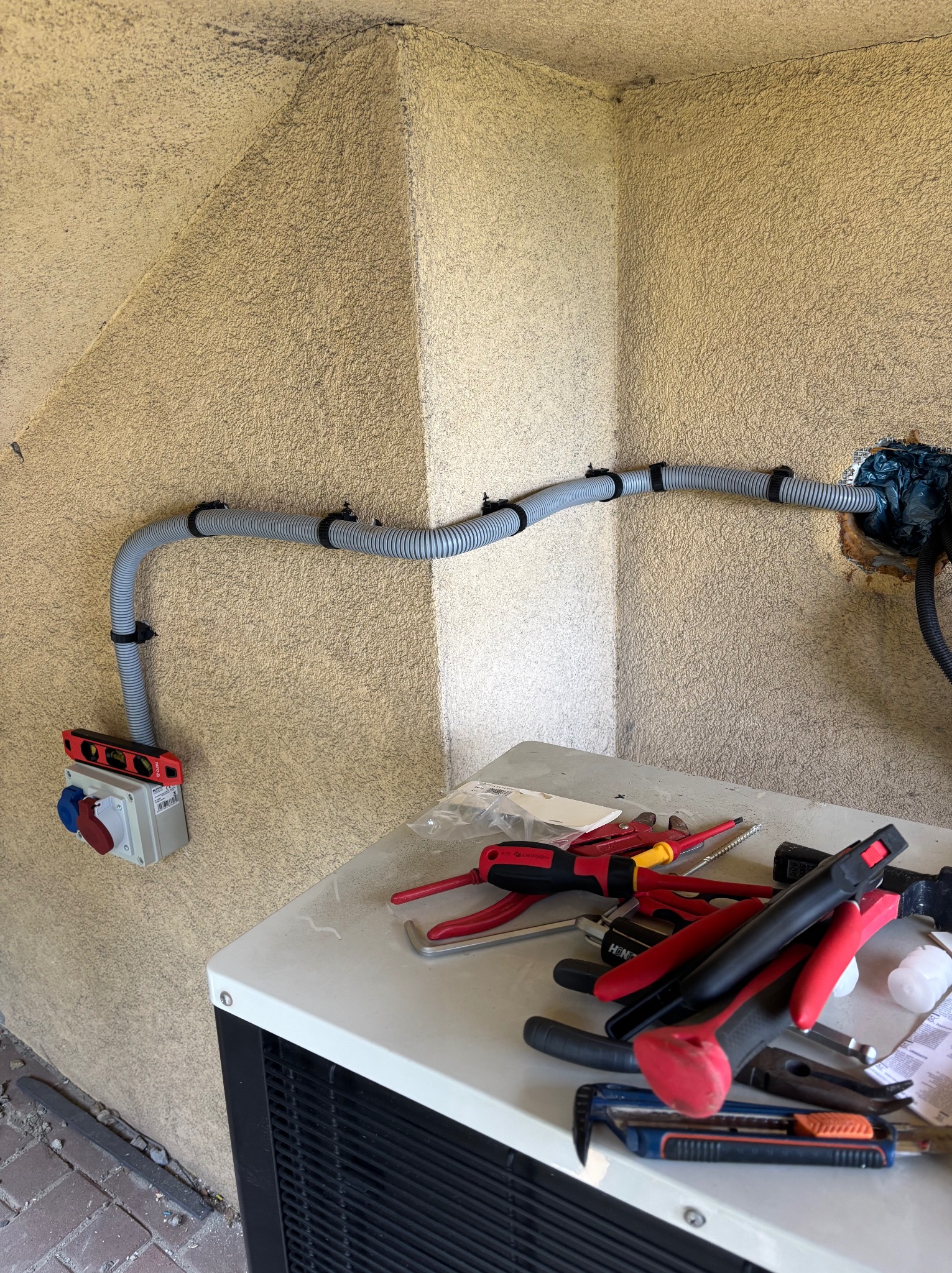Szara, karbowana rura elektryczna zamocowana do ściany z wypustkami, w tle narzędzia na klimatyzatorze. Widoczna puszka elektryczna z czerwono-niebieskim gniazdem.