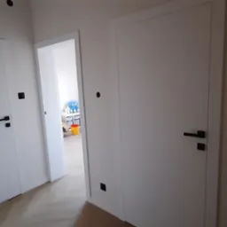 Nowoczesne białe drzwi wewnętrzne z czarną klamką i minimalistyczną ościeżnicą, widok z korytarza na pokój dziecięcy z zabawkami w tle.