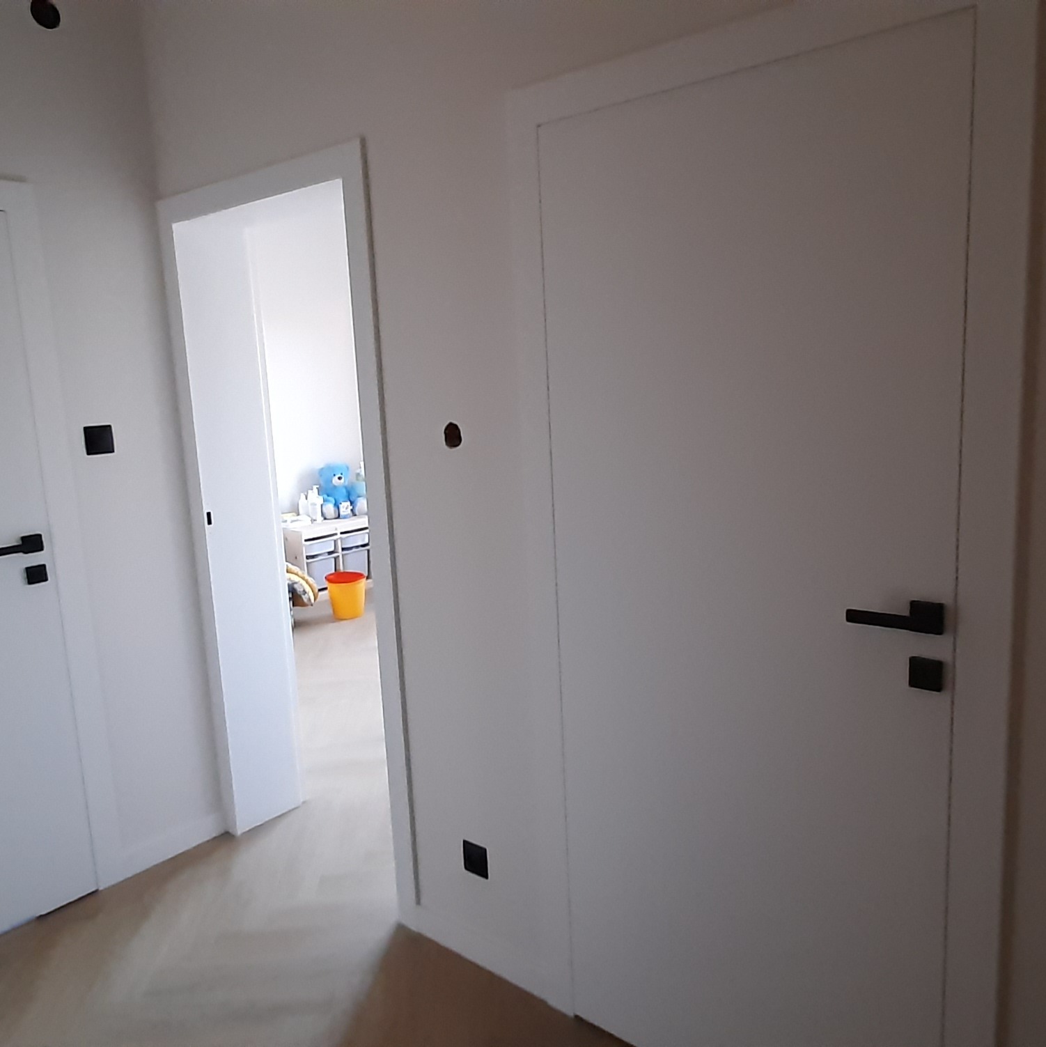 Nowoczesne białe drzwi wewnętrzne z czarną klamką i minimalistyczną ościeżnicą, widok z korytarza na pokój dziecięcy z zabawkami w tle.