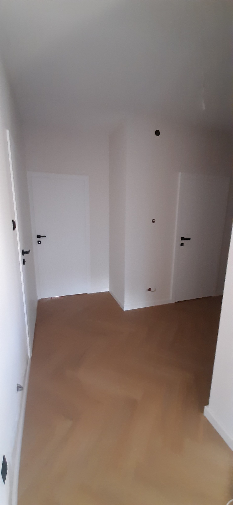Wnętrze korytarza z dwiema białymi drzwiami z czarnymi klamkami i jasnobrązową podłogą, widoczne niedoskonałości na ścianach i podłodze.
