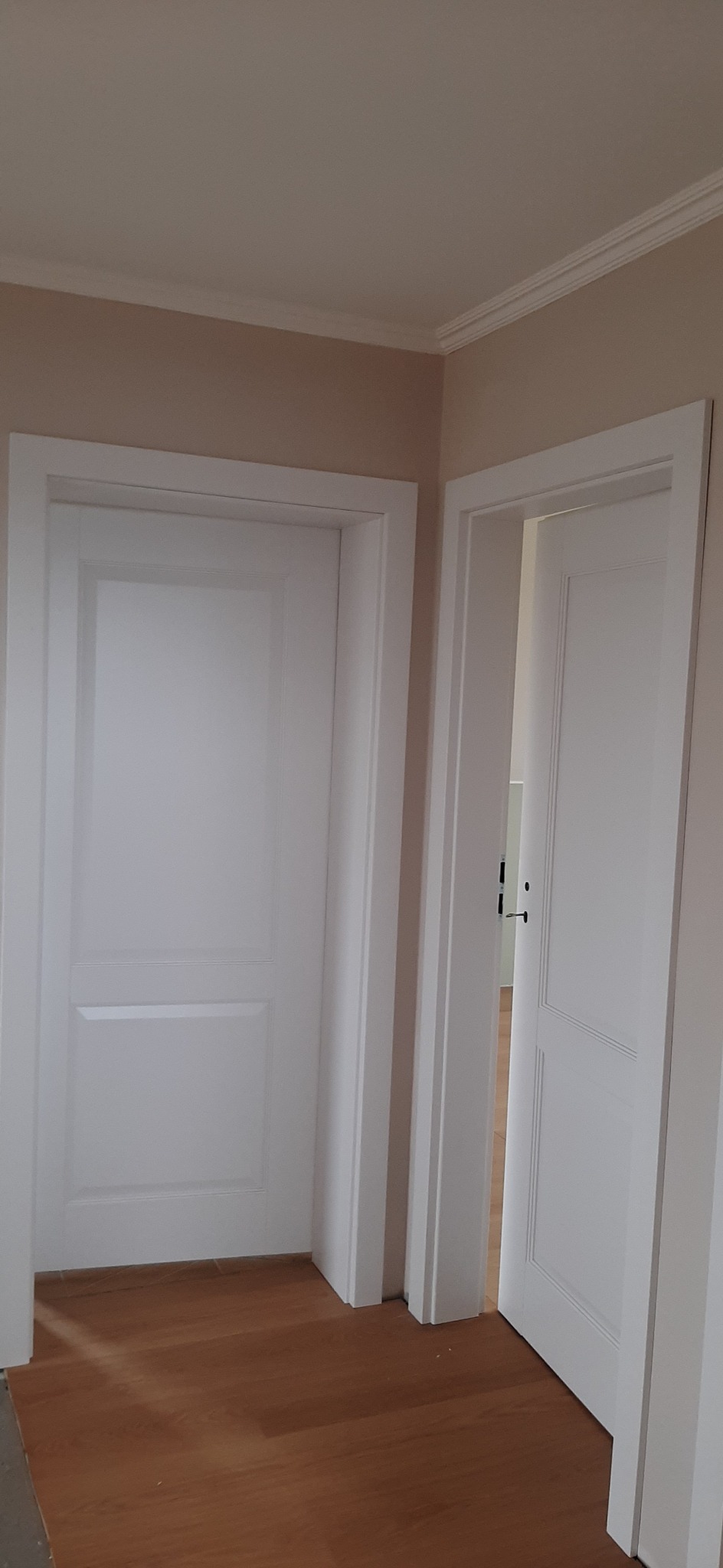 Dwa białe, panelowe drzwi z ościeżnicami, jedno zamknięte, drugie uchylone, na podłodze z paneli w odcieniach brązu.