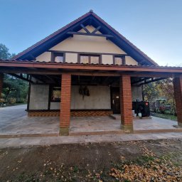 Robert Kromp - Zadaszona wiata z ceglanymi filarami i kostką brukową na podłożu, widok na ogród z drzewami w jesiennych barwach.