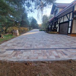 Robert Kromp - Ukończona nawierzchnia z kostki brukowej w odcieniach brązu i szarości, z liniowym odpływem wody, prowadząca do domu z elementami szachulcowymi, pies w tle.