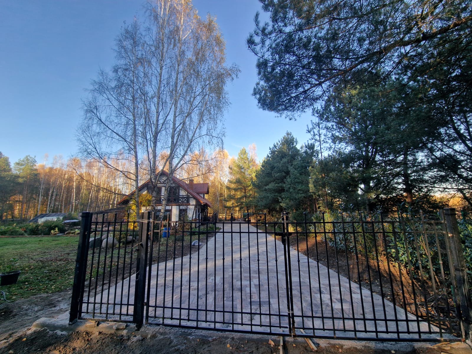 Nowoczesne, czarne ogrodzenie panelowe przed domem z podjazdem wyłożonym kostką, otoczone drzewami w jesiennej scenerii.