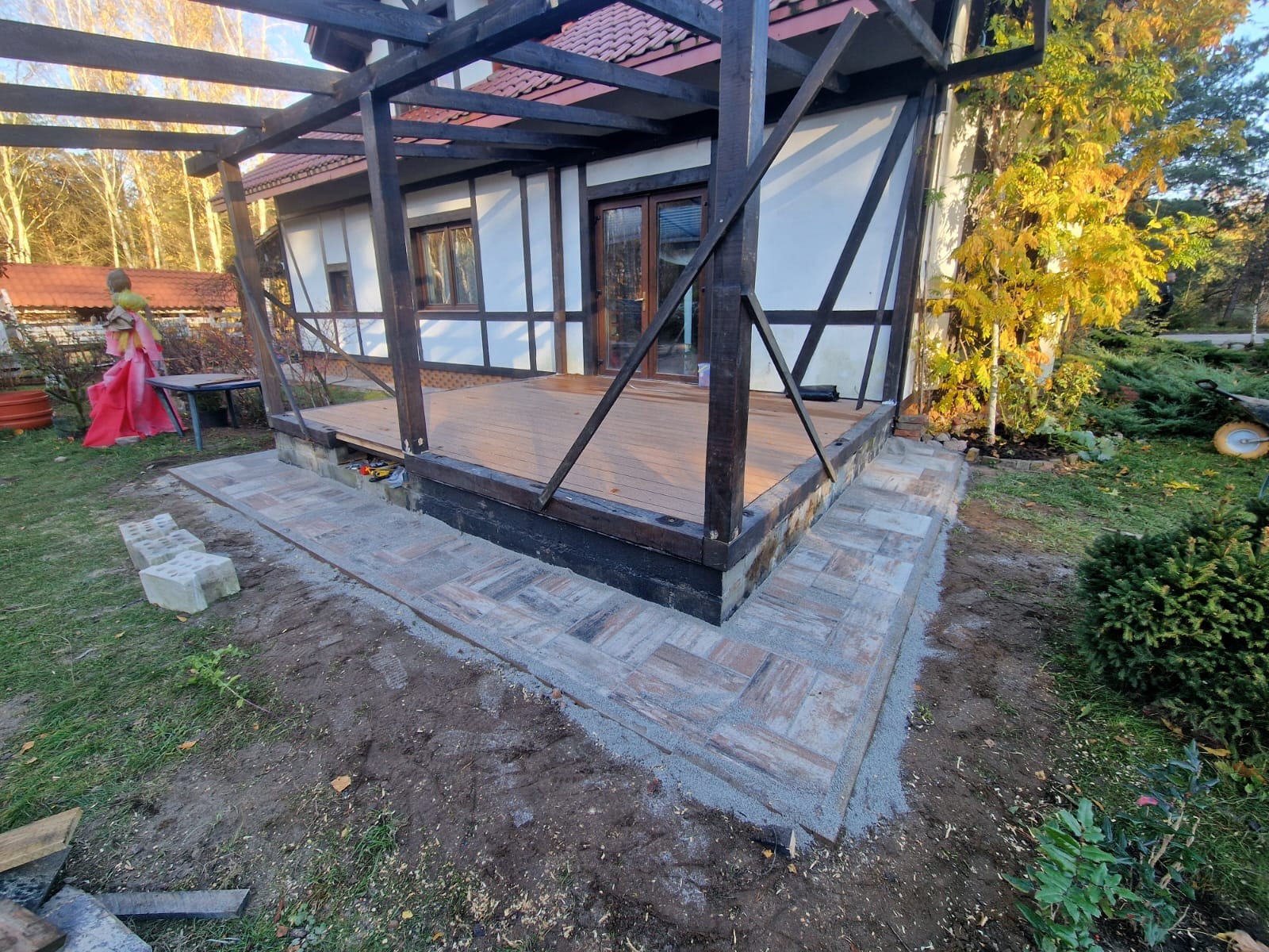 Nowo wyłożony chodnik z kostki brukowej prowadzący do drewnianego tarasu z pergolą przy domu w stylu szachulcowym, widoczne niedokończone prace ziemne.