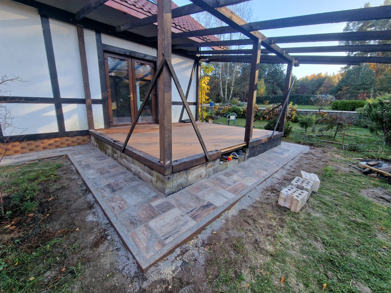 Budowa drewnianej pergoli przy domu z wyłożonym obrzeżem z kostki brukowej. Widoczne narzędzia i materiały budowlane.