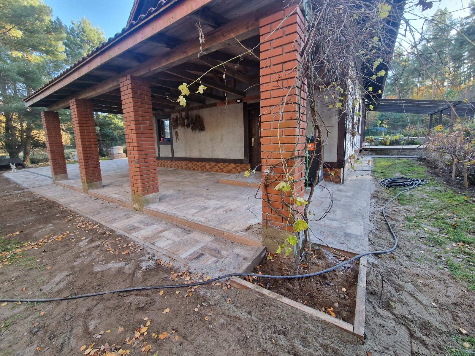 Zadaszony taras z ceglanymi filarami i nową nawierzchnią z kostki brukowej w odcieniach beżu i brązu, widoczne przygotowania do nasadzeń roślin przy filarze.