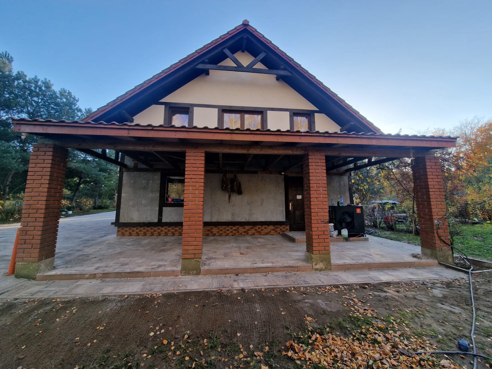 Zadaszona wiata z ceglanymi filarami i kostką brukową na podłożu, widok na ogród z drzewami w jesiennych barwach.