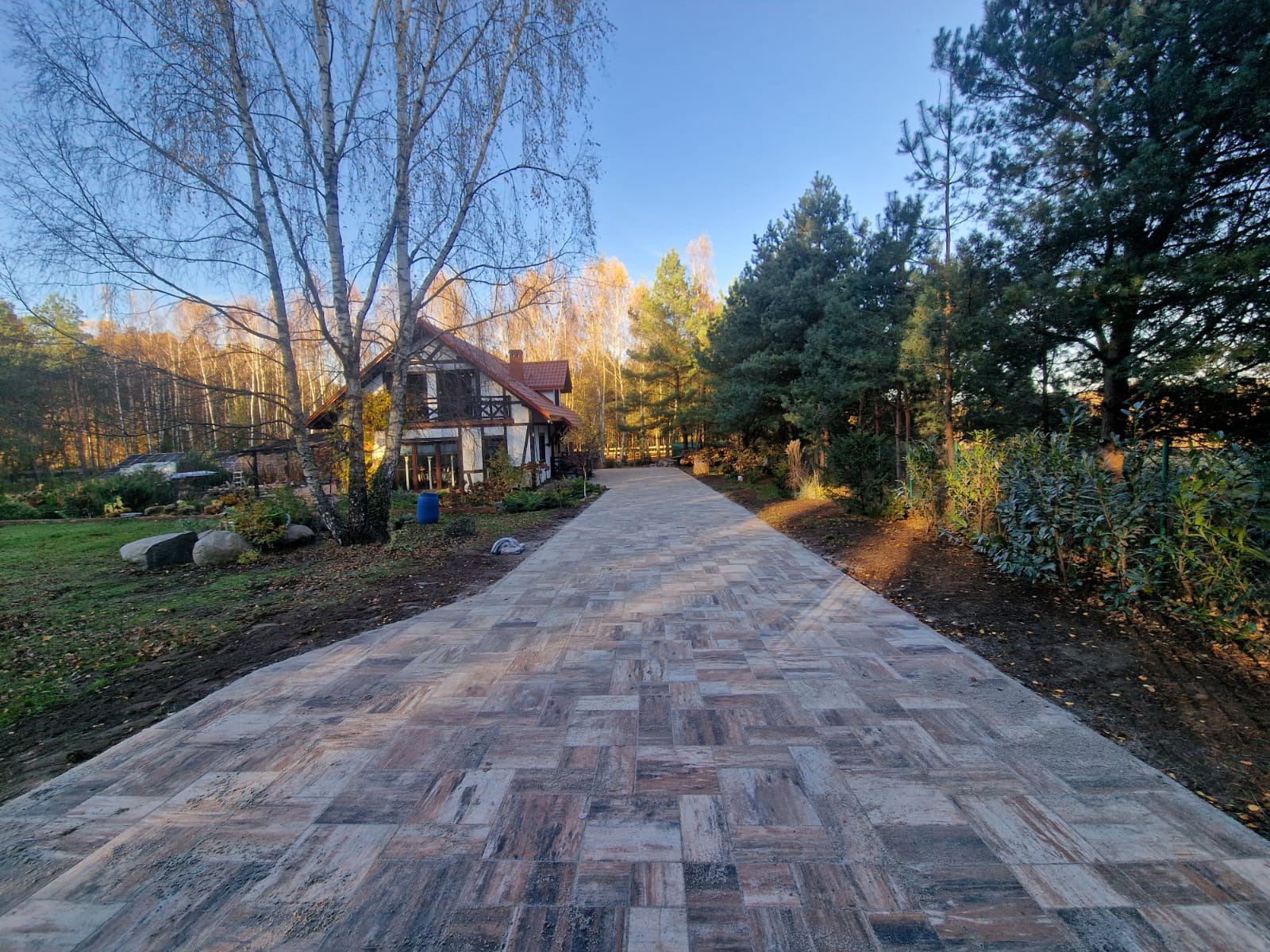 Długa, nowo ułożona droga z kostki brukowej w odcieniach brązu i szarości prowadząca do malowniczego domu w stylu rustykalnym z czerwonym dachem, otoczonego drzewami w jesiennych barwach.