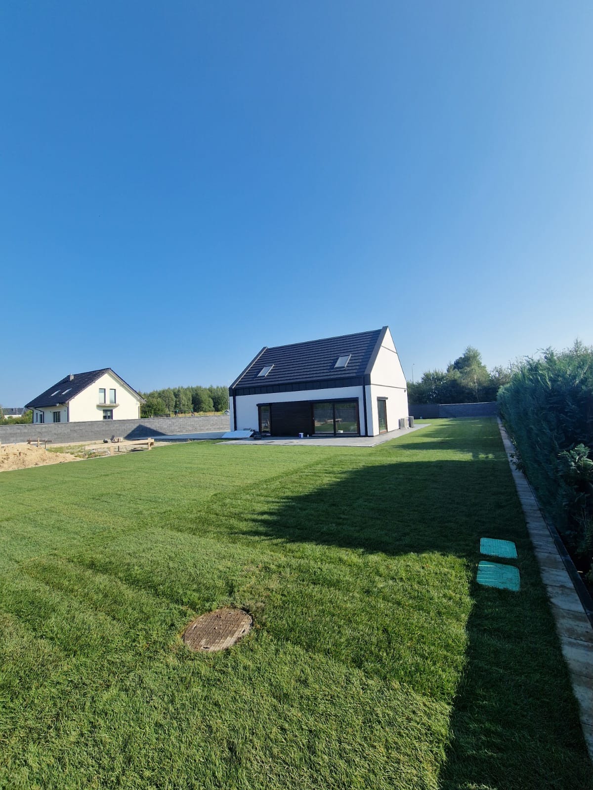 Nowoczesny dom jednorodzinny z czarnym dachem i dużymi oknami, otoczony świeżo założonym trawnikiem i tujami, widoczny właz do studzienki kanalizacyjnej w trawie.