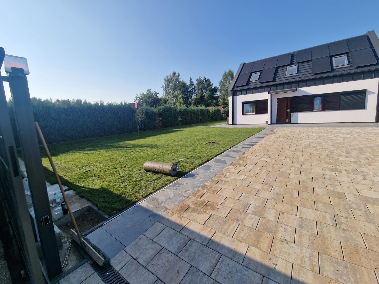 Nowoczesny dom z beżową kostką brukową na podjeździe, świeżo rozłożony trawnik z rolki i zielony żywopłot w tle.