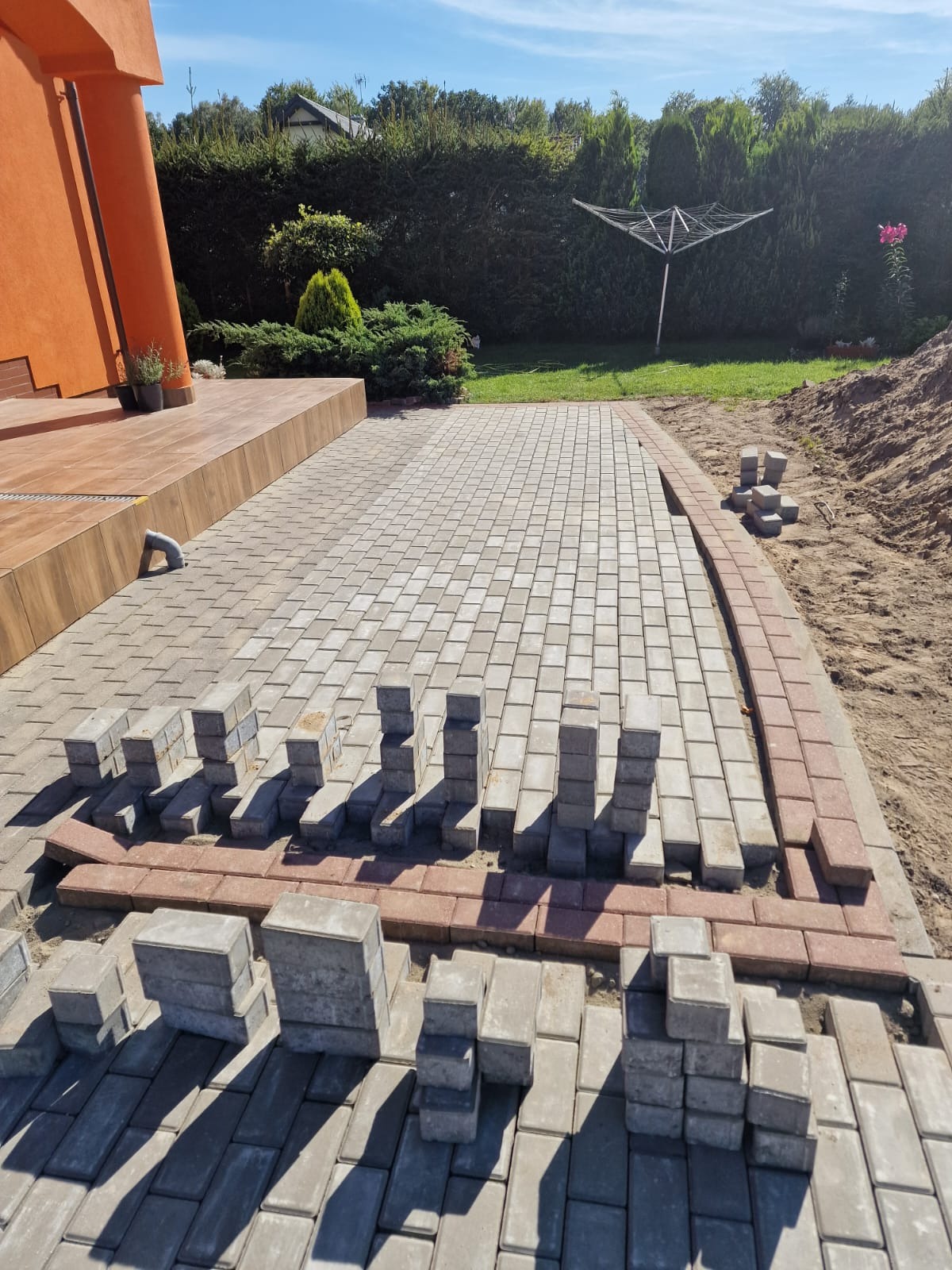 Fragment podjazdu w trakcie budowy z szarej kostki brukowej i bordowej obwódki, ułożone stosy kostek, widok na dom z pomarańczową elewacją i zielony ogród.