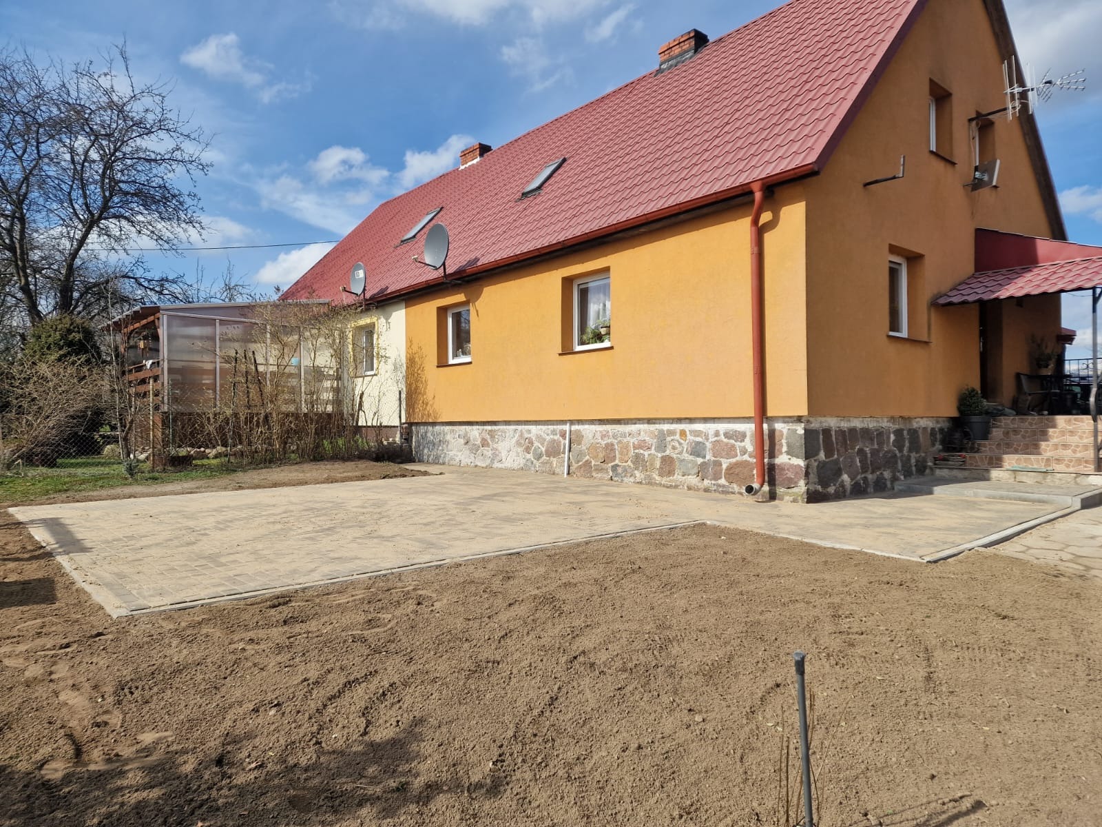 Ułożony plac z kostki brukowej przed domem z żółtą elewacją i czerwonym dachem, widoczny przygotowany teren pod trawnik.