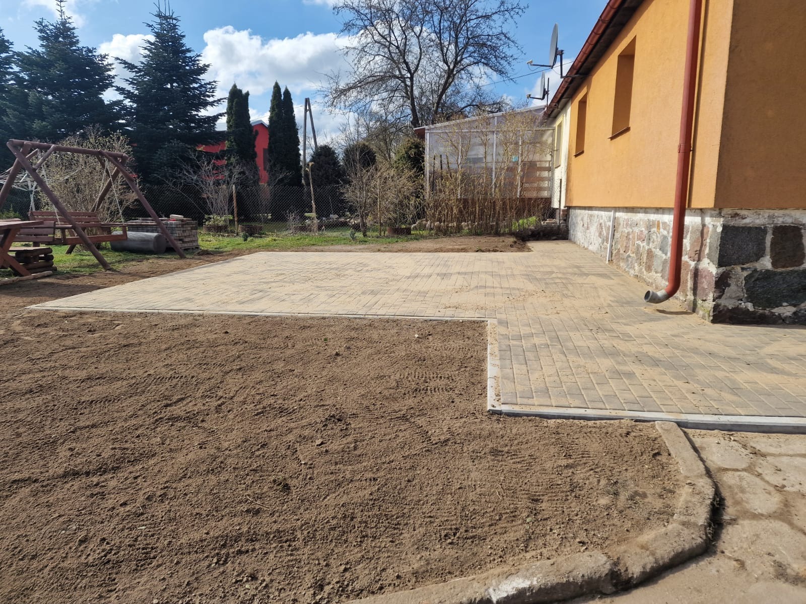 Fragment posesji z częściowo ułożoną beżową kostką brukową, widoczna przygotowana podłoże z ziemi, w tle dom z pomarańczową elewacją i elementy ogrodu.