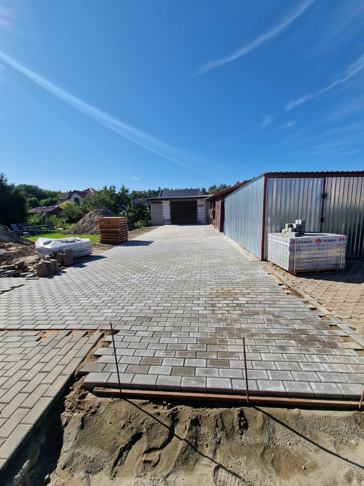 Świeżo ułożona szara kostka brukowa prowadząca do garażu blaszanego, widoczne palety z kostką i przygotowane podłoże z piasku.