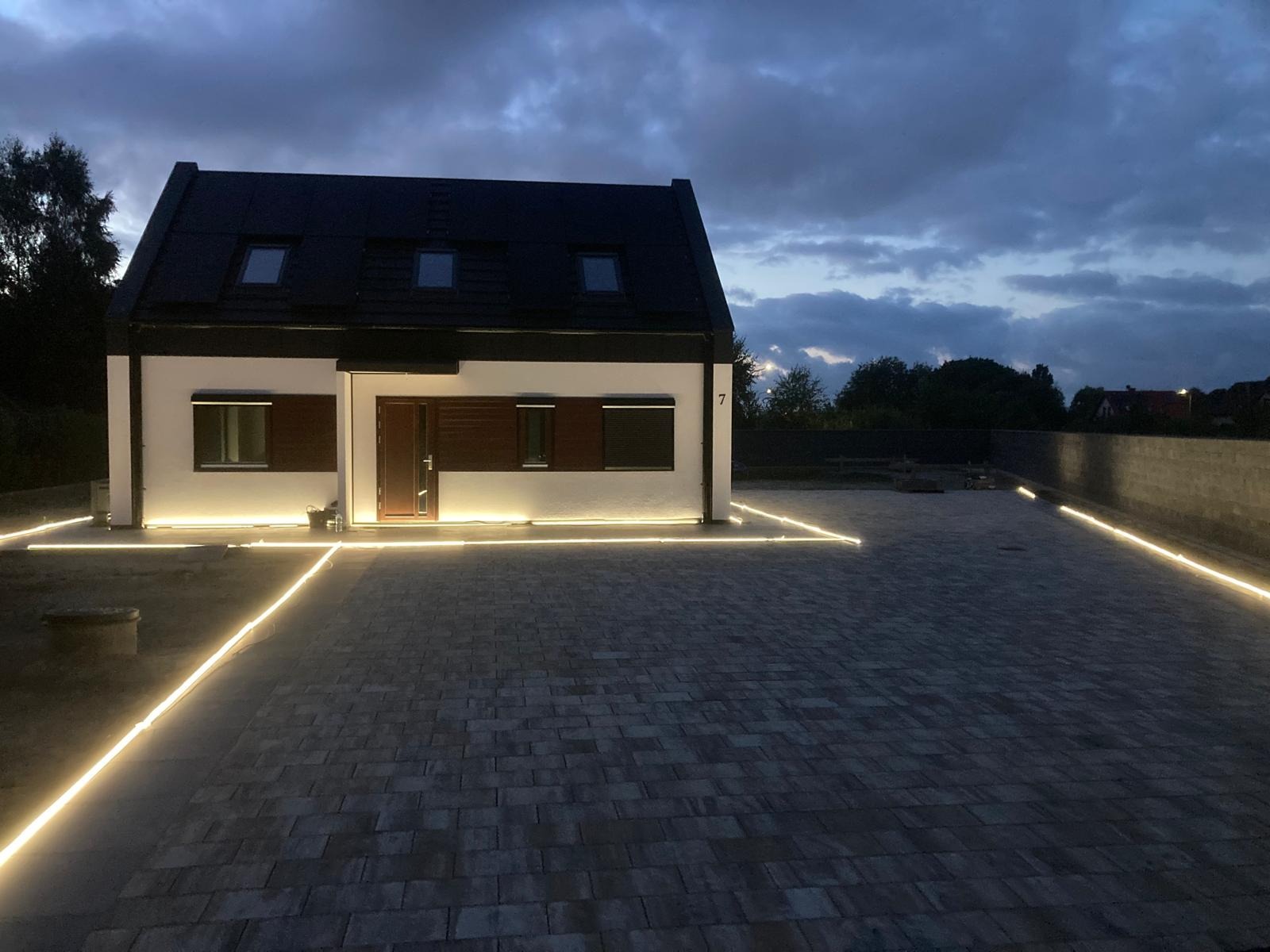 Nowoczesny dom jednorodzinny z podświetloną elewacją i brukowanym podjazdem o zmierzchu, widoczne linie świetlne wzdłuż kostki i budynku.